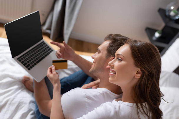 selective focus of cheerful woman holding credit card near man using laptop with blank screen in bedroom  - Foto, Imagen