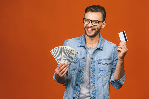 Εικόνα του συγκλονισμένος νεαρός όμορφος γενειοφόρος άνδρας θέτει απομονωμένη πάνω πορτοκαλί τοίχο φόντο κρατώντας χρήματα και πιστωτική κάρτα κάνουν χειρονομία νικητής. - Φωτογραφία, εικόνα