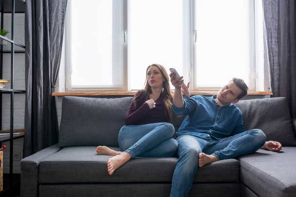 woman sitting on sofa near man holding remote controller from air conditioner while feeling hot  - Photo, Image