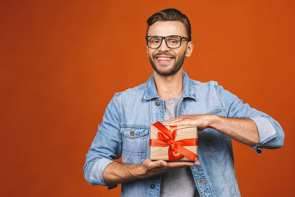 С праздником! Очаровательная фотография привлекательного мужчины с красивой улыбкой с настоящей коробкой, изолированной над оранжевой стеной
.  - Фото, изображение
