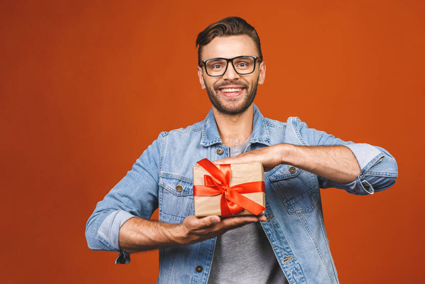 С праздником! Очаровательная фотография привлекательного мужчины с красивой улыбкой с настоящей коробкой, изолированной над оранжевой стеной
.  - Фото, изображение