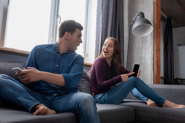 excited girl pointing with finger at smartphone with blank screen and looking at boyfriend in living room  - Foto, Imagen