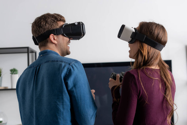 KYIV, UKRAINE - MAY 15, 2020: cheerful couple in virtual reality headsets playing video game in living room  - Foto, Imagen