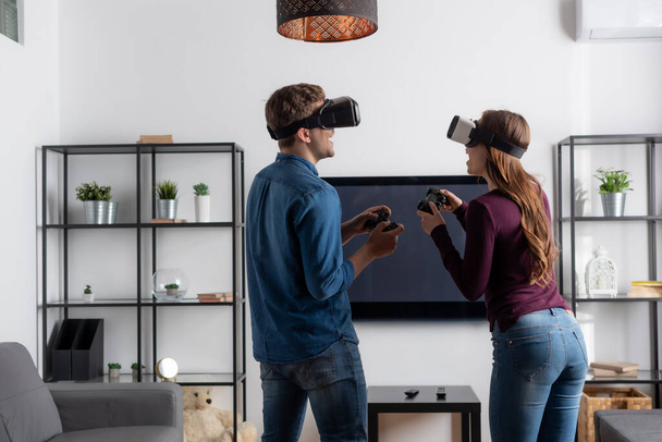 KYIV, UKRAINE - MAY 15, 2020: cheerful couple in virtual reality headsets holding joysticks and playing video game in living room  - Photo, Image