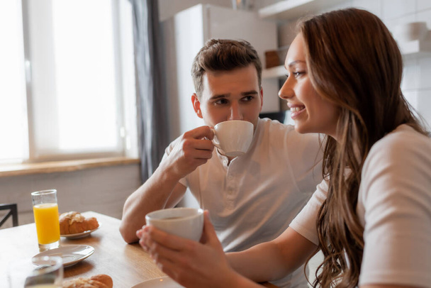 selective focus of man drinking coffee and looking at cheerful girlfriend in kitchen  - Fotografie, Obrázek