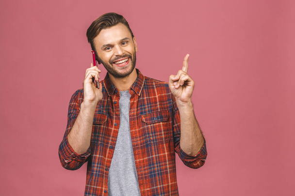 Casual young man speaking on the phone and pointing aside while smiling to the camera. Isolated on pink.  - Photo, Image