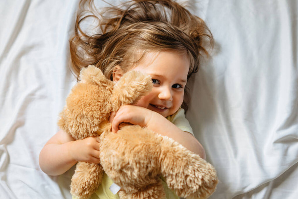 Χαμογελαστό κοριτσάκι που κρατάει αρκουδάκι και ξαπλώνει στο κρεβάτι με λευκά σεντόνια στο σπίτι. Ευτυχισμένο παιδί. Άνω όψη.  - Φωτογραφία, εικόνα