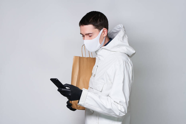 Homem usando uma máscara protetora para evitar a propagação do Coronavirus segurar saco de compras e se comunica via smartphone
. - Foto, Imagem
