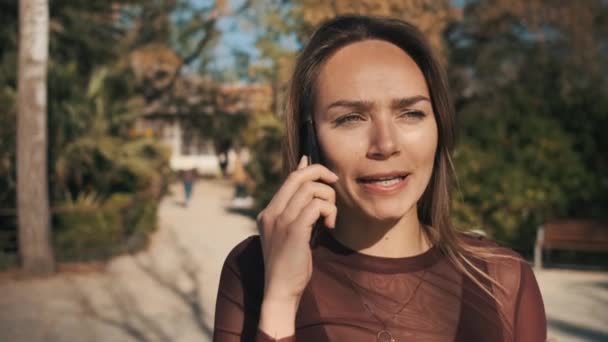 Tracking shot annoyed and disappointed beautiful girl arguing with boyfriend emotionally talking on the phone in city park - Video