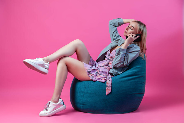 Blond meisje zit in een blauwe tas stoel in een korte jurk met een halslijn en benen gekruist op een roze achtergrond met een telefoon in haar hand en houdt haar hoofd vast. Thuis werken in de zelfisolatiemodus. - Foto, afbeelding