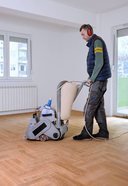 Sanding hardwood floor with the grinding machine. Repair in the apartment. Carpenter doing parquet wood floor polishing maintenance work by grinding machine - Photo, Image