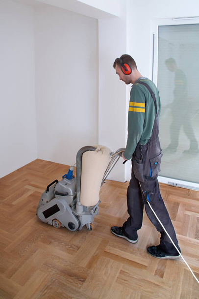 Lijado piso de madera dura con la máquina de pulir. Reparación en el apartamento. Carpintero haciendo trabajos de mantenimiento de pulido de suelo de madera de parquet mediante rectificadora
                                - Foto, Imagen