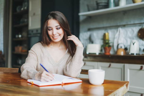 Uśmiechnięta brunetka azjatycki młoda kobieta w dorywczo robi notatki w dziennej książce z filiżanką herbaty na kuchni w domu - Zdjęcie, obraz