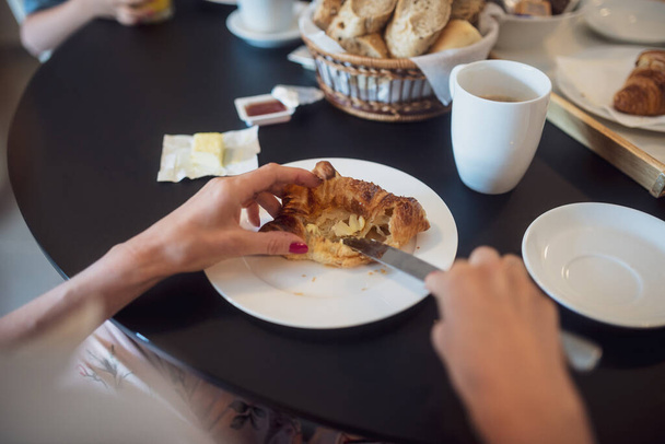 Vrouwelijke handen verspreiden boter en jam op heerlijke croissant op volledige ontbijttafel. - Foto, afbeelding