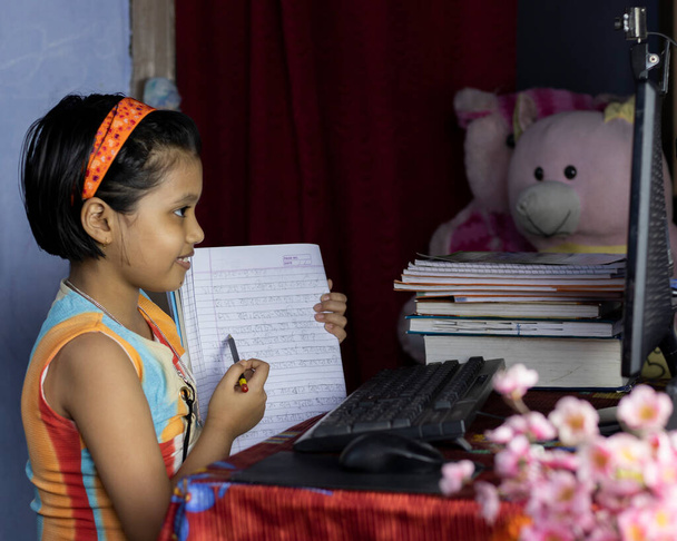 una bambina indiana con un volto sorridente che frequenta la classe online sul computer desktop e mostra il taccuino all'insegnante durante l'epidemia di pandemia del COVID-19
 - Foto, immagini
