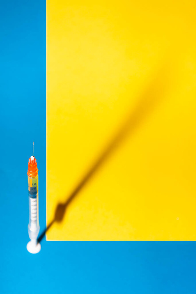 Injekční stříkačka pro vakcínu nebo inzulín s jejím stínem na modrém a žlutém pozadí. Přímé nebo selektivní zaměření. Zdravotní koncepce, vakcinace, koronární onemocnění nebo léky - Fotografie, Obrázek