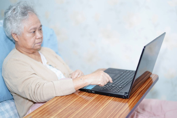 Asian senior or elderly old lady woman patient using notebook computer work from home while sitting on bed in nursing hospital ward. - Photo, Image