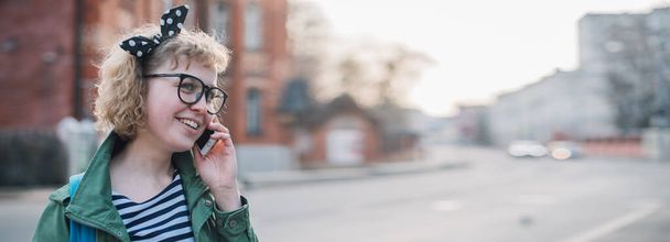 Молодая женщина разговаривает по телефону на европейской улице. Стильная блондинка в очках и полосатая рубашка для прогулки по старому городу в солнечный день
 - Фото, изображение