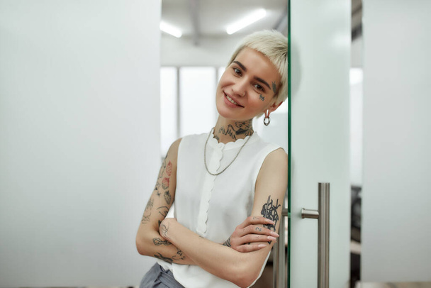 Being happy. Young, smiling beautiful blonde tattooed businesswoman leaning on the glass door, keeping arms crossed and looking at camera while standing in modern office - Φωτογραφία, εικόνα