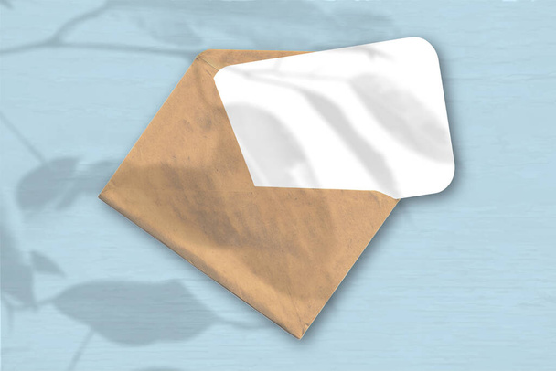 Um envelope com uma folha de papel branco texturizado sobre o fundo azul. Mockup com uma sobreposição de sombras de fábrica. A luz natural lança sombras de uma fábrica exótica. Orientação horizontal
 - Foto, Imagem