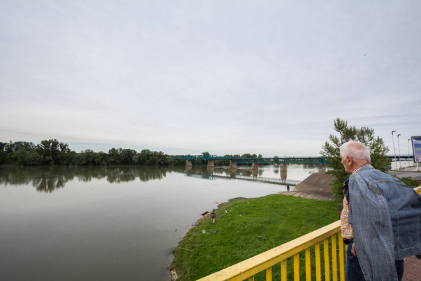 BRCKO, BOSNIA - MAY 6, 2017: Old Senior man looking at a Steel bridge crossing the Sava river at the border between Bosnia and and Croatia, an official border crossing of the European Union (EU) - Photo, Image