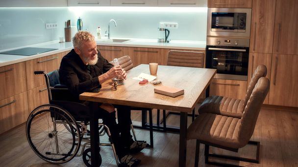 Frustration. Senior disabled man in wheelchair lighting cannabis in the bowl of glass water pipe or bong in kitchen. Marijuana tools on the table. Legalization and dependence on light drugs concept - Foto, imagen