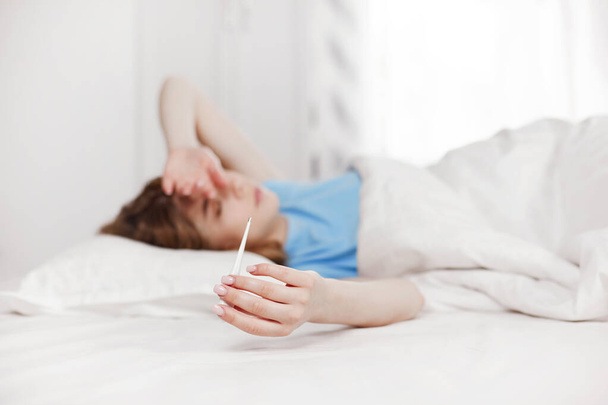 A beautiful girl during the illness lies on the bed and takes her temperature. Poor health during a fever. Blurred background, space for text - Fotoğraf, Görsel