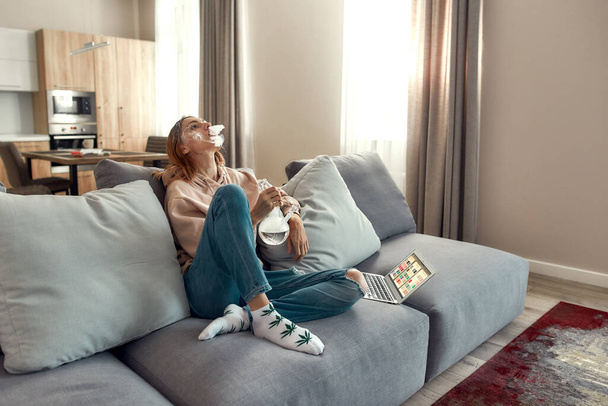 Relax and Enjoy. Young caucasian woman exhaling the smoke while smoking marijuana from a bong or glass water pipe, sitting on the couch. Cannabis and weed legalization concept - Фото, зображення