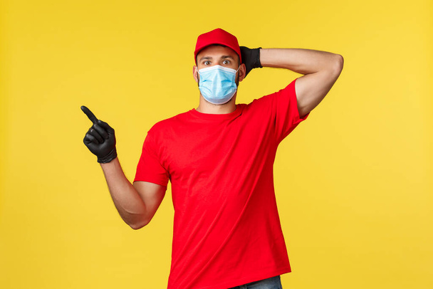 Express delivery during pandemic, covid-19, safe shipping, online shopping concept. Impressed courier in red logistic company uniform, stare and point left excited, yellow background - 写真・画像