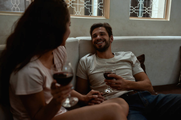 Romance never goes out of fashion. Happy couple relaxing, drinking wine and talking while sitting on couch at home - Foto, imagen