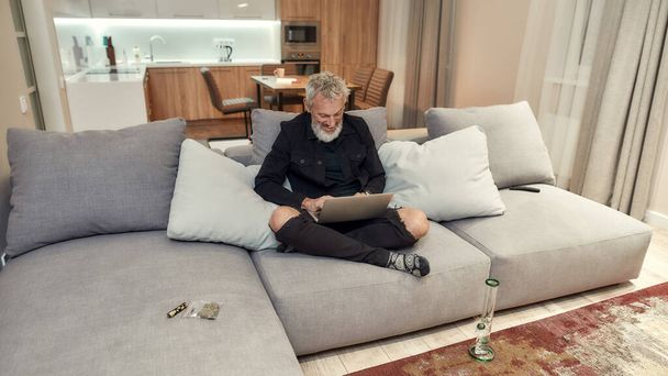 Working. Bearded middle-aged man using laptop while sitting on the couch. Bong or glass water pipe, buds in plastic bag and lighter near him. Cannabis and weed legalization concept - Photo, Image