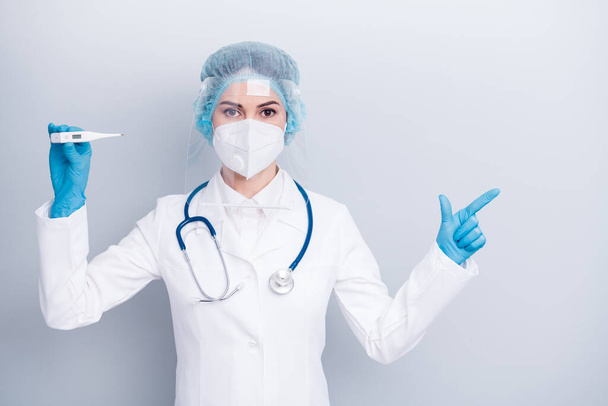 Photo of disease virologist lady hold thermometer direct finger side advising make vaccination wear coat respirator mask facial plastic protection surgical hat gloves isolated grey background - 写真・画像