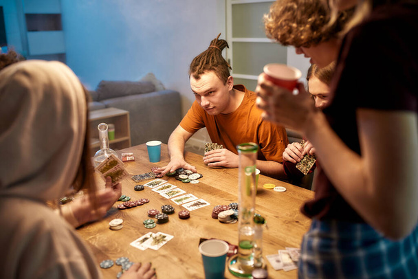 Going all in. Young people playing poker after smoking marijuana at home. Marijuana tools, bong on the table. Home party - Foto, Imagen