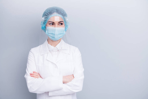 Photo of attractive confident virologist doctor lady arms crossed good mood look side empty space wear medical lab coat mask facial plastic protection surgical cap isolated grey background - Фото, зображення