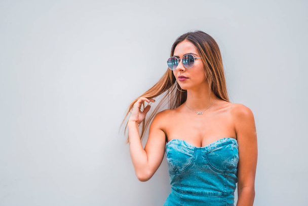 Lifestyle session, a young caucasian brunette in a blue denim dress and sunglasses on a plain gray background, with room for copy and paste, looking to the left - Foto, afbeelding