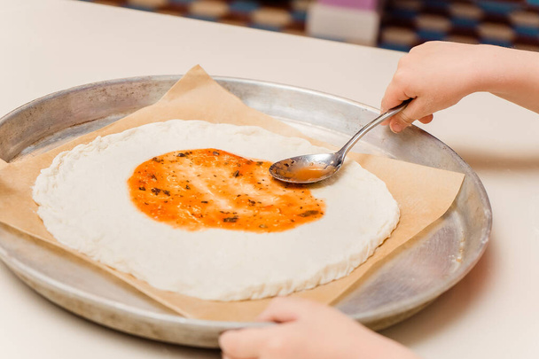 El niño cocina. La mano pone una cucharada de salsa en la base de la pizza. Clase culinaria para niños sobre hornear
 - Foto, imagen
