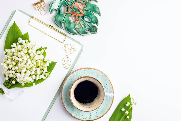 Home office desk table with coffee cup, bouquet of lilies of the valley flat lay. Workplace with notebook and decorations on white background. Blogging, freelance, feminine, florist concept. Top view - Photo, Image