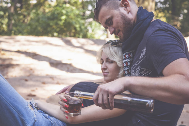 Couple of young people sitting in park and drinking alcohol. Alcoholism and alcohol addiction people concept. - Фото, изображение