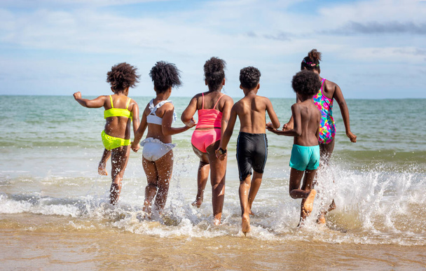 A gyerekek homokon futkároznak a parton, egy csapat gyerek fogja egymás kezét a parton nyáron, visszapillantva a tengerre és a kék égre. - Fotó, kép