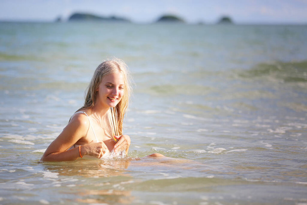 Mulher sorridente de biquíni sentada na praia tropical, mulher feliz de roupa de banho de cor branca desfrutando de férias de verão deitada na areia branca contra o céu azul. Férias, férias, conceito de estilo de vida. Espaço de cópia - Foto, Imagem