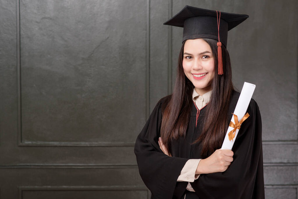 Portrait of young woman in graduation gown smiling and cheering on black background  - Фото, изображение