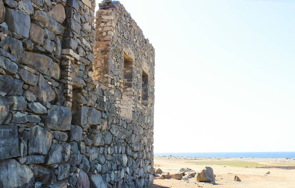 De oude ruïnes van een groot gebouw staan op een zandstrand bij het water. Grote kasseien en blauw water zijn zichtbaar. Aruba augustus 2014 - Foto, afbeelding
