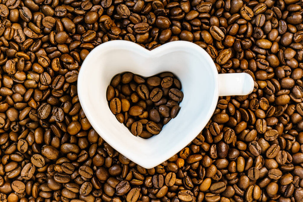 Coffee time. White cup in shape of heart on roasted coffee beans background. Top view - Valokuva, kuva