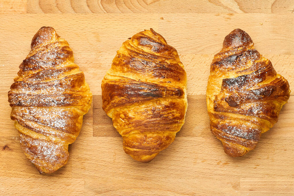 Vista superior de tres croissants recién horneados con polvo de azúcar en un escritorio de madera. Pastelería francesa o italiana
 - Foto, Imagen