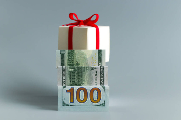 A hundred dollars folded like a ladder and a gift box tied with red ribbon. The concept of hoarding, wealth and surprises - Photo, Image