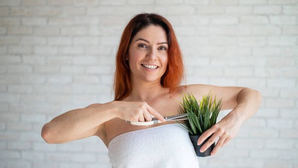 A smiling red-haired woman in a white terry towel cuts a green plant in a pot with scissors. Imitation of armpit depilation. Hair removal concept. Procedure for hair removal of unwanted vegetation. - Foto, imagen
