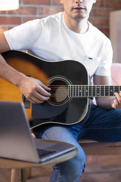 Young man sits in a room on an armchair, learns to play guitar online. Online education, online music lessons, online vocal and guitar training. Stay home. - Photo, image