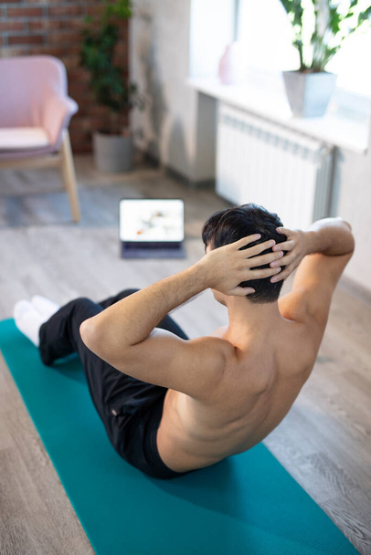 Young slim man training at home. Workout online fitness lessons on a mat with a laptop, doing russian twisting and sit-ups. Online training, online fitness, stay home, quarantine, online training. - Zdjęcie, obraz