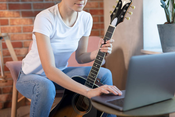 Young woman sits in a room on an armchair, learns to play guitar online. Online education, online music lessons, online vocal and guitar training. Stay home. - Photo, image