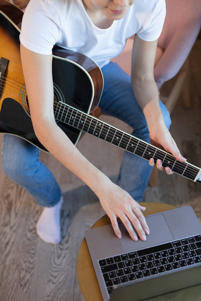 Young woman sits in a room on an armchair, learns to play guitar online. Online education, online music lessons, online vocal and guitar training. Stay home. - Фото, изображение